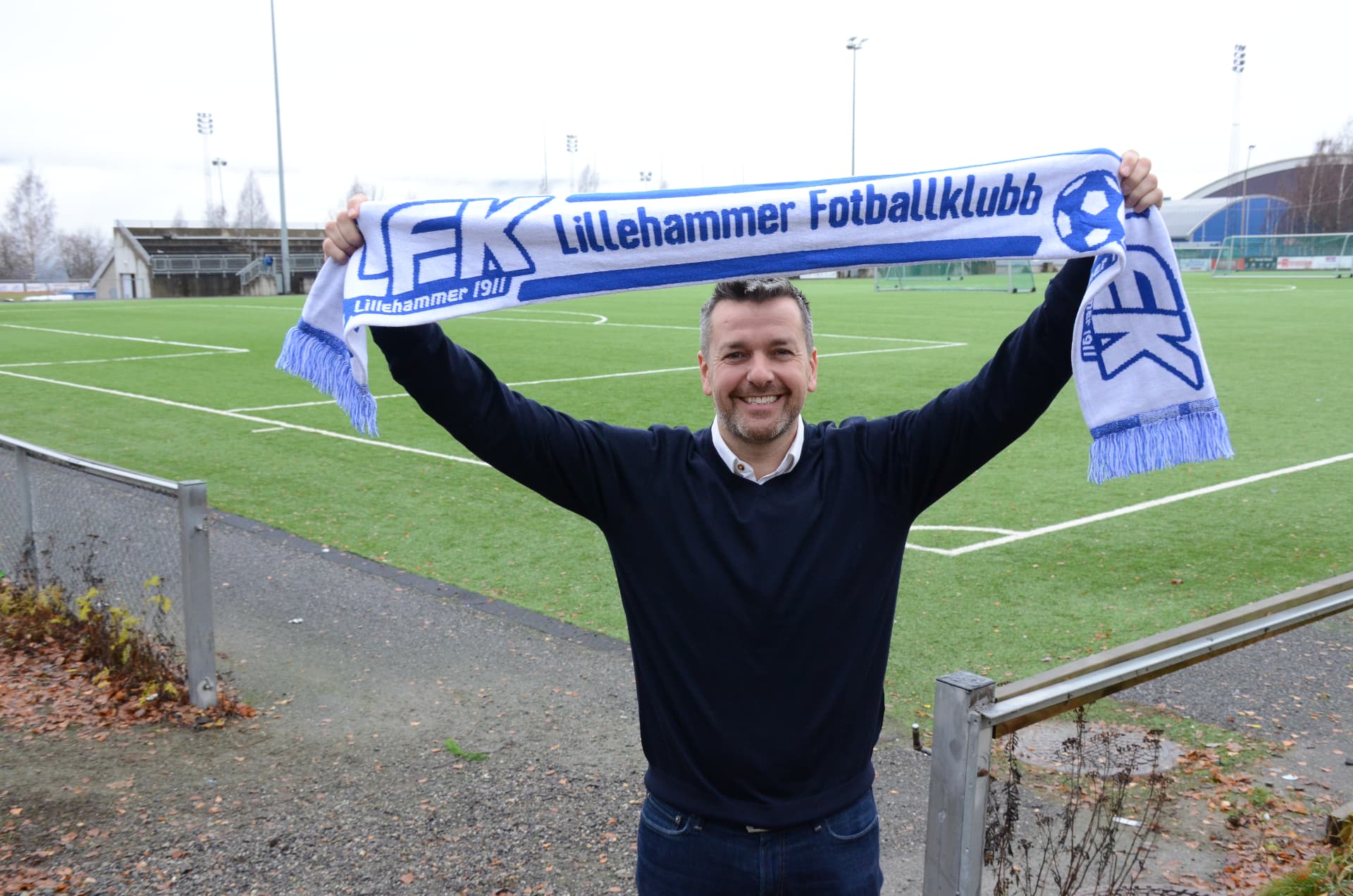 Russell Rusty Hullett holding a Lillehammer FK scarf beside the football pitch
